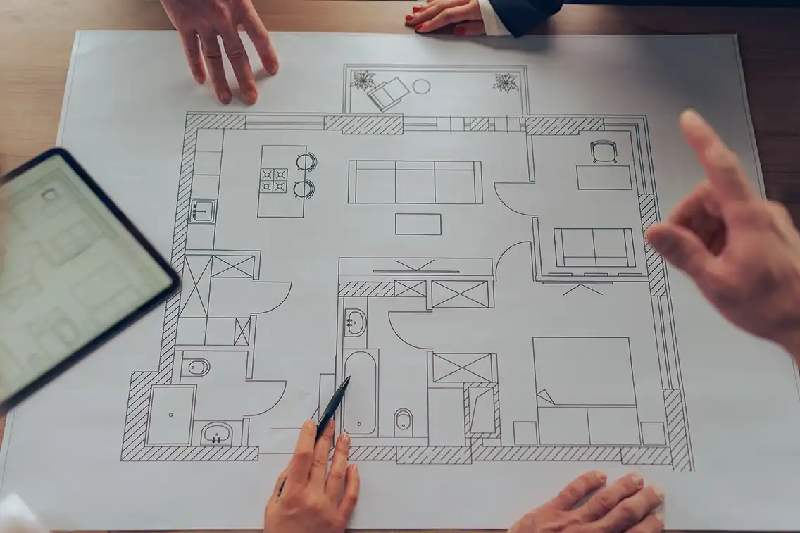 Collaborative planning session with people examining a detailed floor plan, including rooms and furniture, using both paper and digital tools.