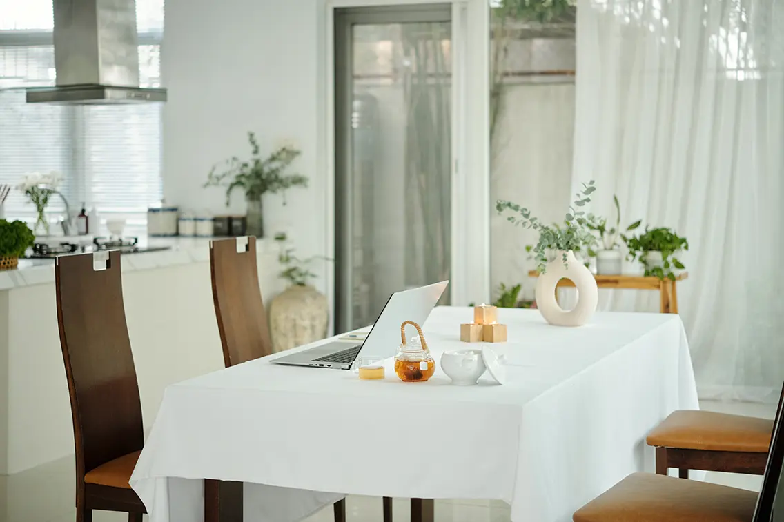 Modern open kitchen opening to a dining room. The room has white themed decoration. Beside the dining table the green plants add charm to the room.