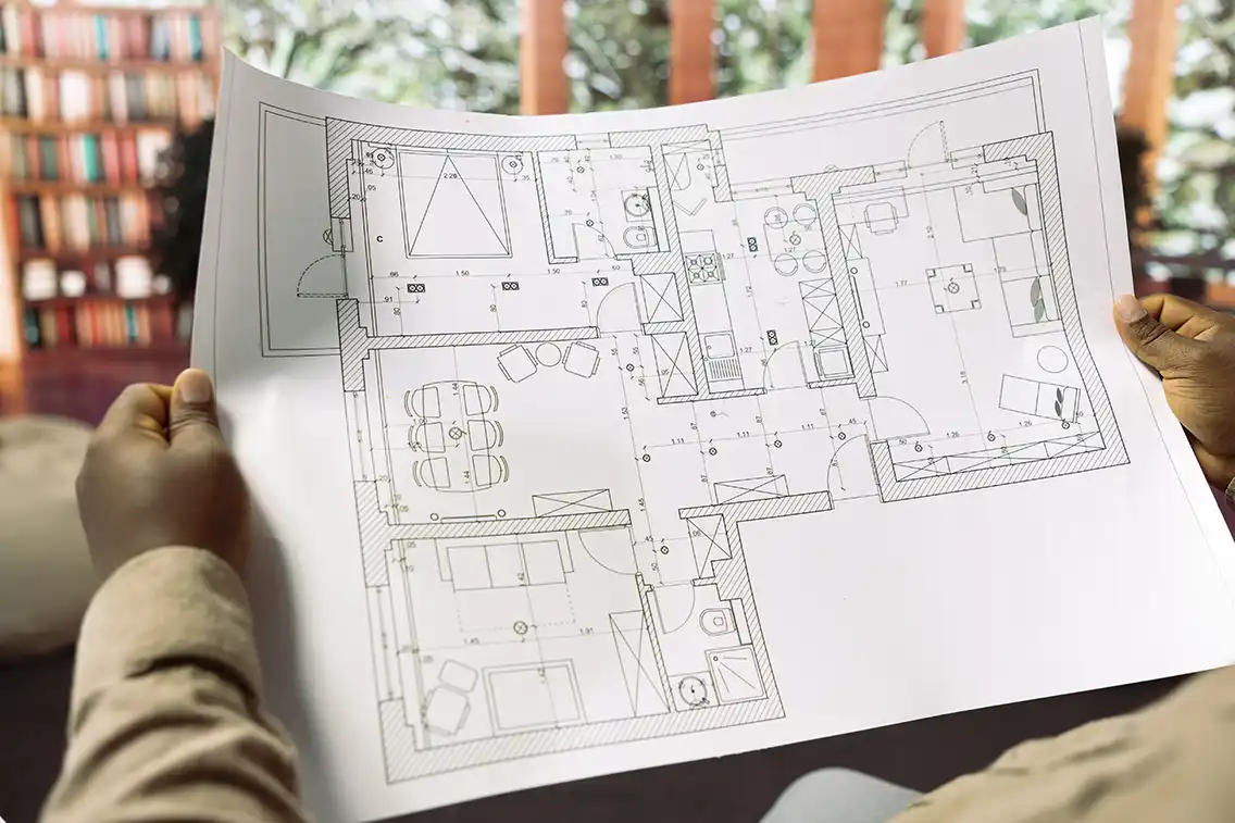 Person holding a detailed architectural floor plan of a house, with rooms, furniture, and measurements. Blurred bookshelf and window in the background.