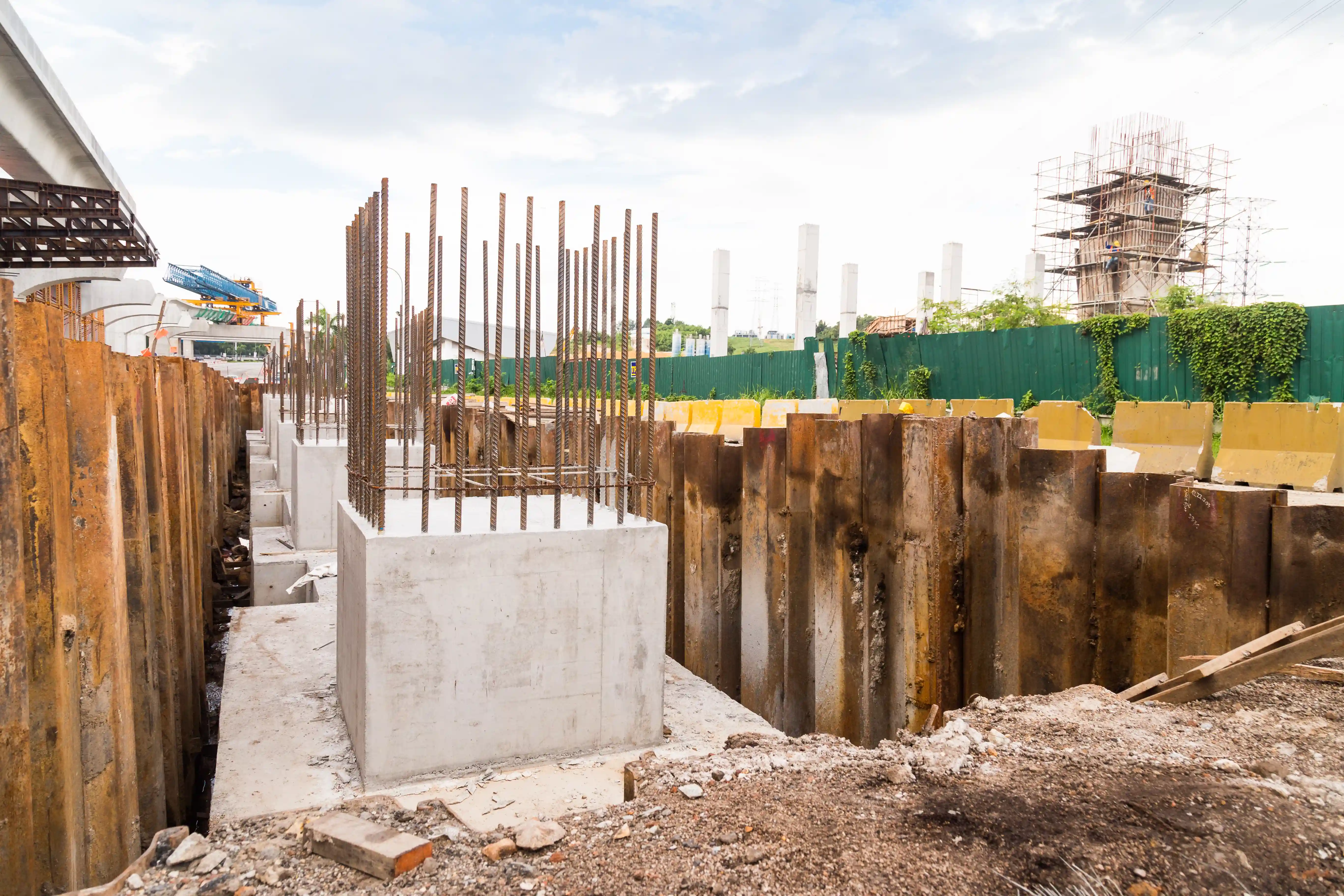 Construction site with several concrete foundations and rebar structures surrounded by steel sheet piles, scaffolding, and additional concrete columns in the background, indicating ongoing building activities.