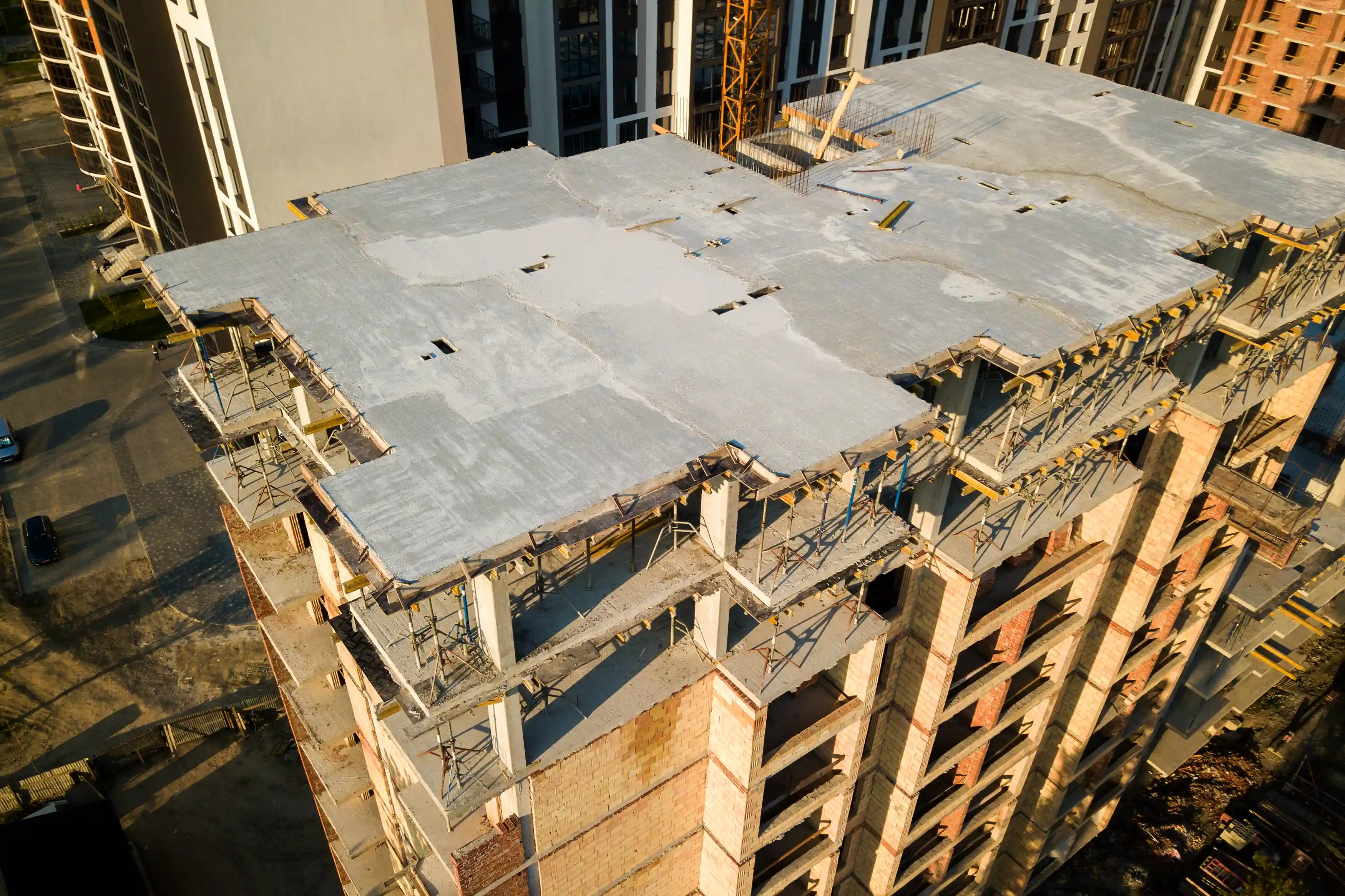 Construction site with a partially built multi-story building, featuring concrete framework, scaffolding, and construction materials. The top floor has a freshly poured concrete slab with rebar protruding.