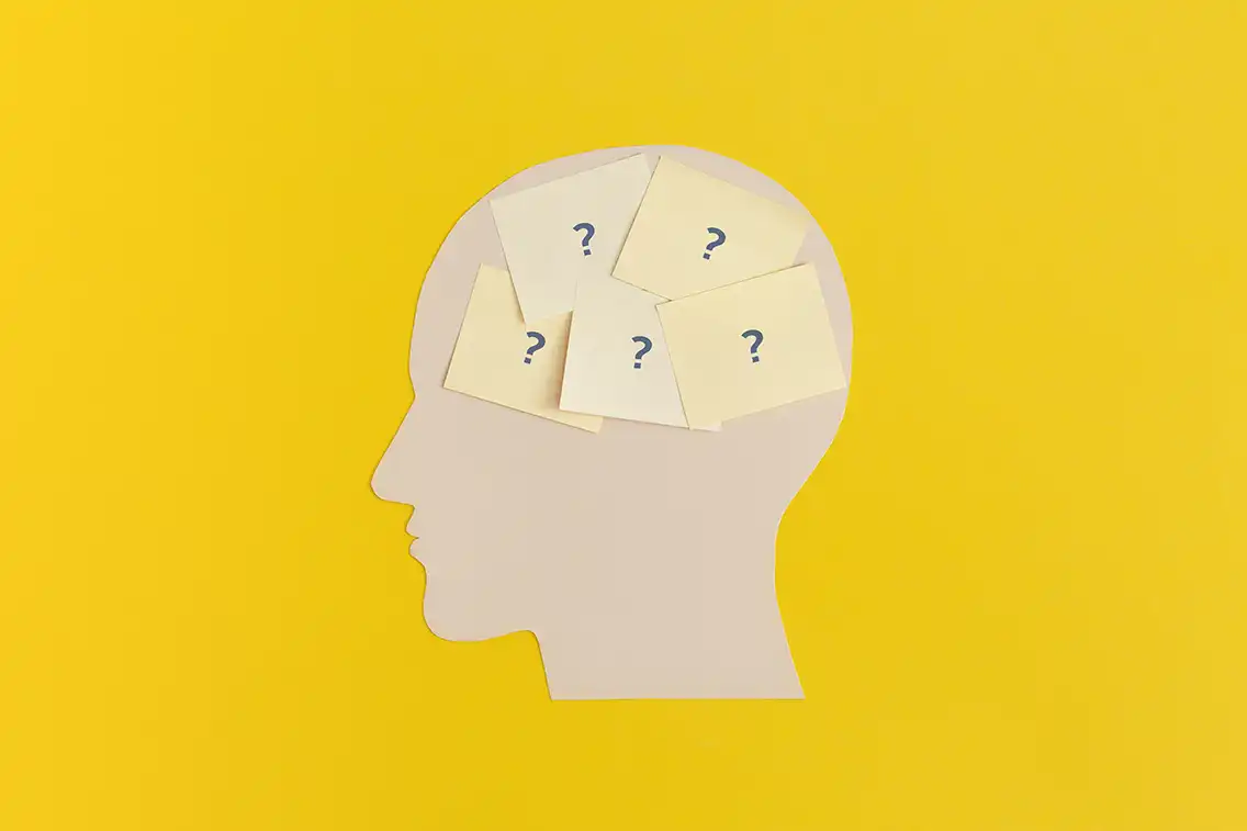 Silhouette of a head filled with sticky notes containing question marks, set on a yellow background, symbolizing confusion, questions, or brainstorming.