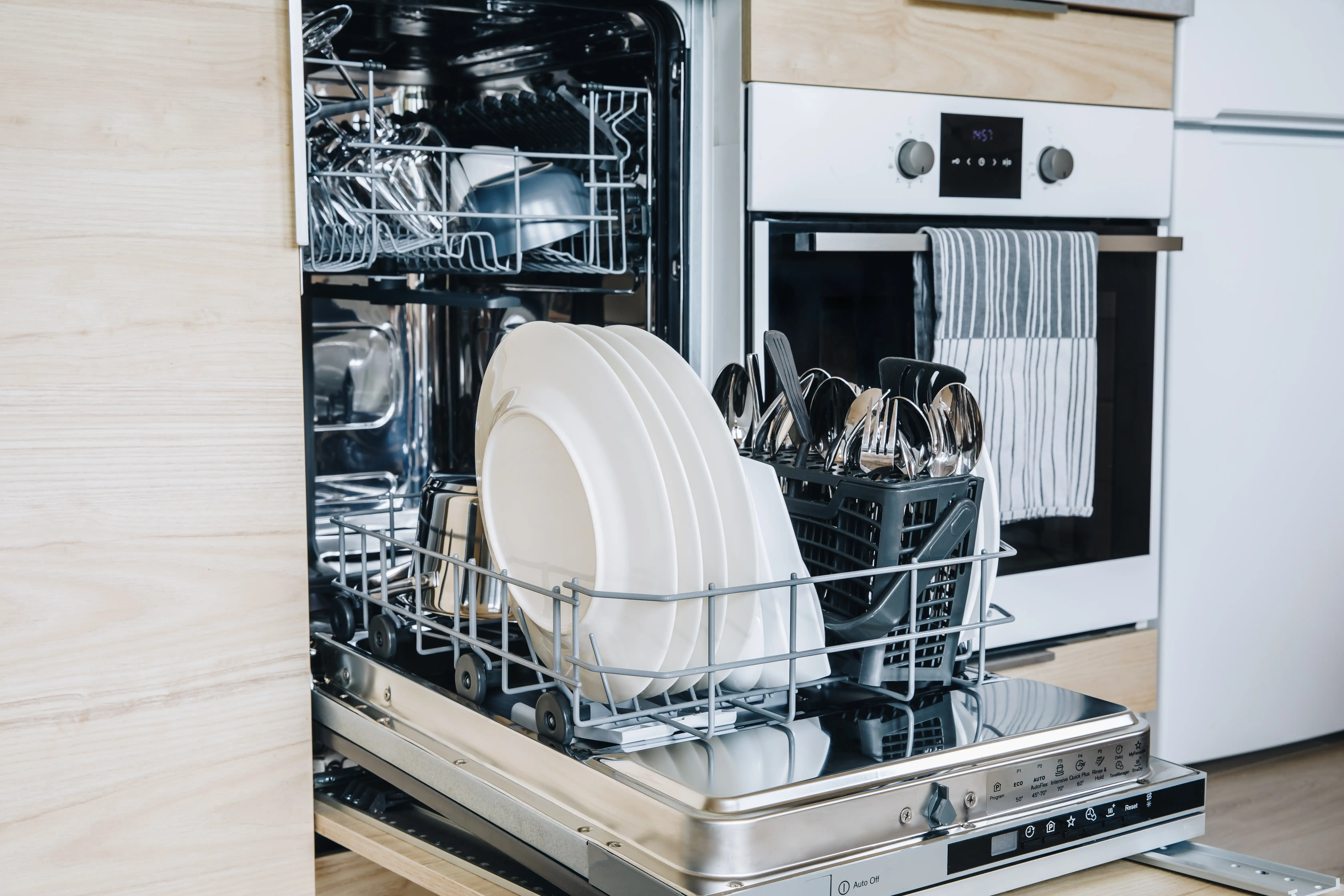 Open dishwasher in a modern kitchen, loaded with clean dishes including plates, bowls, and cutlery. Oven with a striped towel in the background.