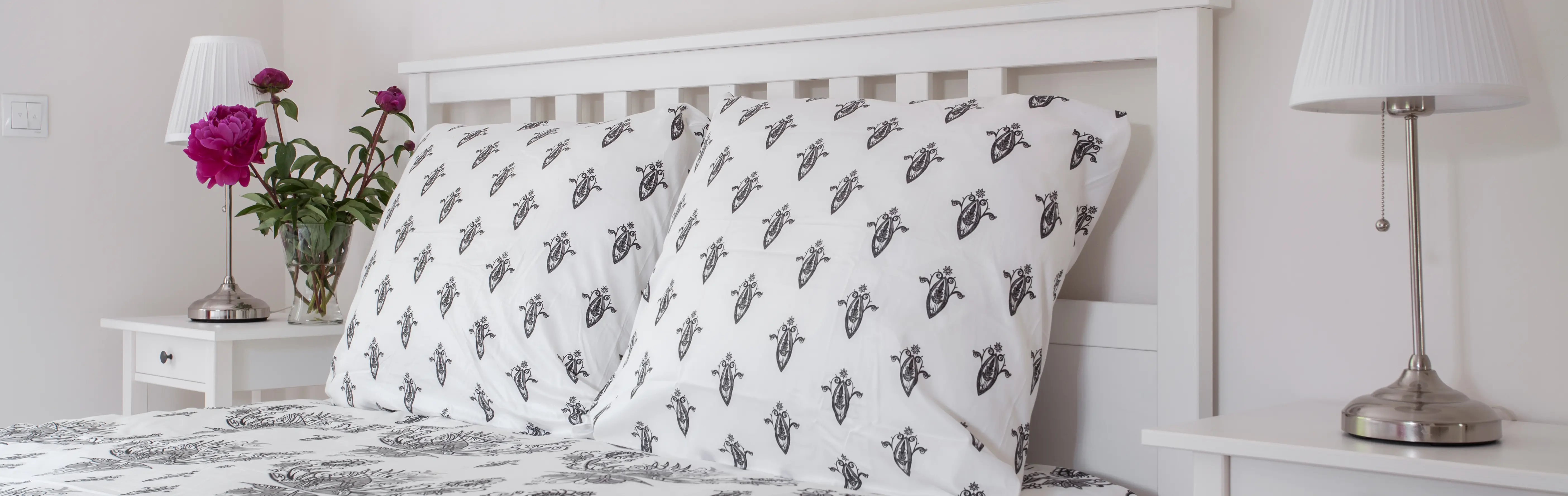A cozy bed that is covered in a printed cotton bedsheet. There are soft pillows. Beside the bed, there are side tables with decors. 