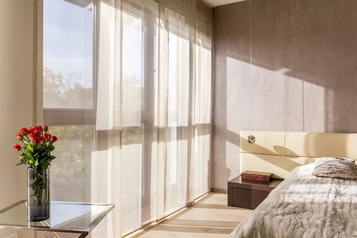 A bright bedroom with sheer curtains covering large windows, natural light streaming in, a vase of red flowers on a glass table, and a cozy bed with white bedding.