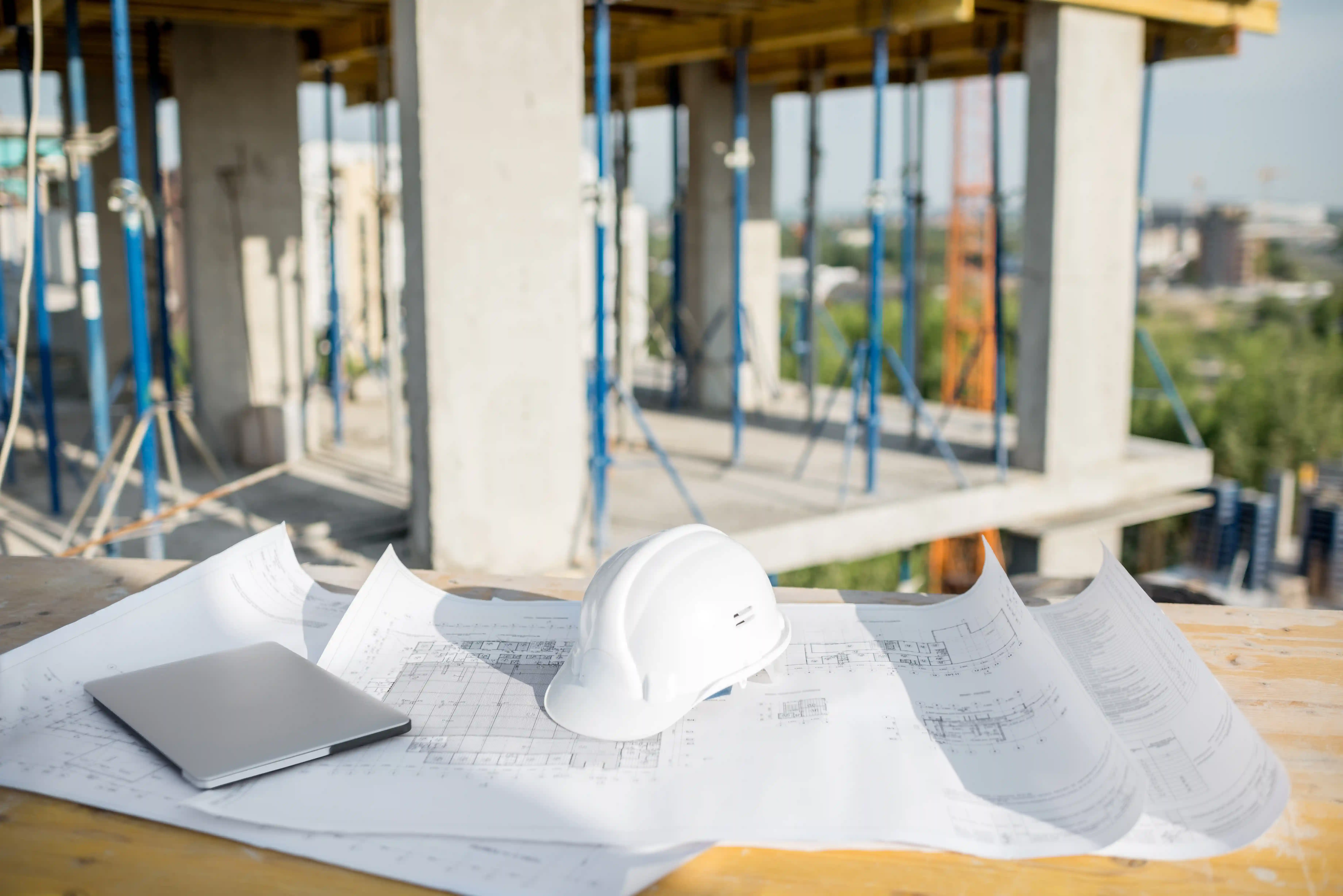 Construction site with a table displaying architectural blueprints, a white hard hat, and a closed laptop, with concrete columns and blue scaffolding poles in the background.