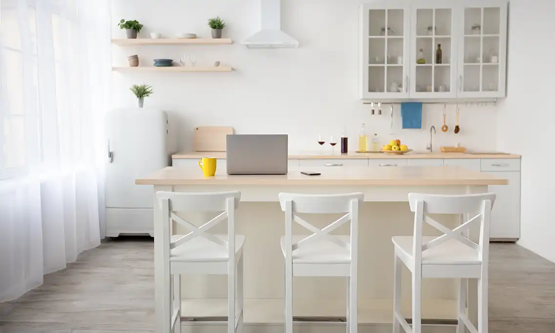 A modern kitchen with a central island, three white chairs, a wooden countertop, a laptop, a yellow mug, and a smartphone. Background includes a white refrigerator, wooden shelves with dishes and plants, a white range hood, and a sink area with various kitchen items.