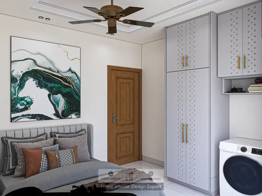 Stylish bedroom with upholstered bed, decorative cushions, abstract wall art, wooden door, white wardrobe, visible washing machine, and ceiling fan enhancing comfort and design.